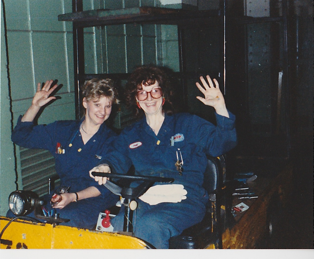 Cheryl Thompson and a coworker in blue coveralls smiling and waving from an industrial tow tractor on the plant floor.